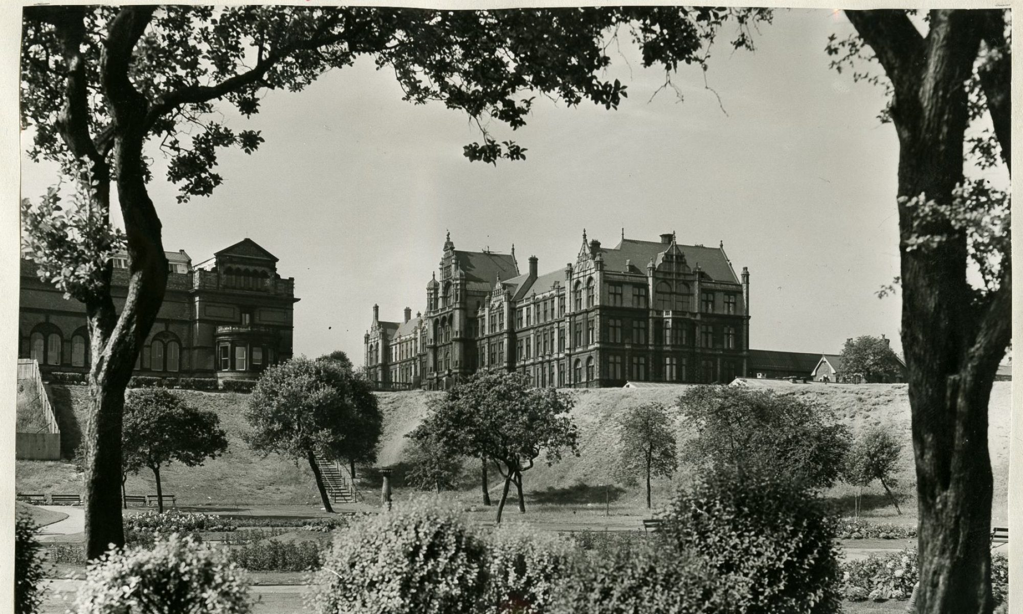 A historic black and white photograph of Peel Park and Peel Building in Salford, England. In the foreground and midground are shrubs and trees filled with leaves. To the left of the image is Salford Museum and Art Gallery, and to the right is Peel Building, a large red brick building with many windows, some overlooking the park.