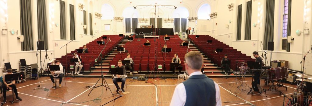 A wide image of the interior of Peel Hall in Salford. Around 200 red seats are at the centre of the image, and a wooden floor in the foreground. There are large white walls with closed dark grey curtains. Around 20 musicians with wind, brass and percussion instruments sit in the space, widely spread out due to covid social distancing. They are rehearsing for a recorded performance.