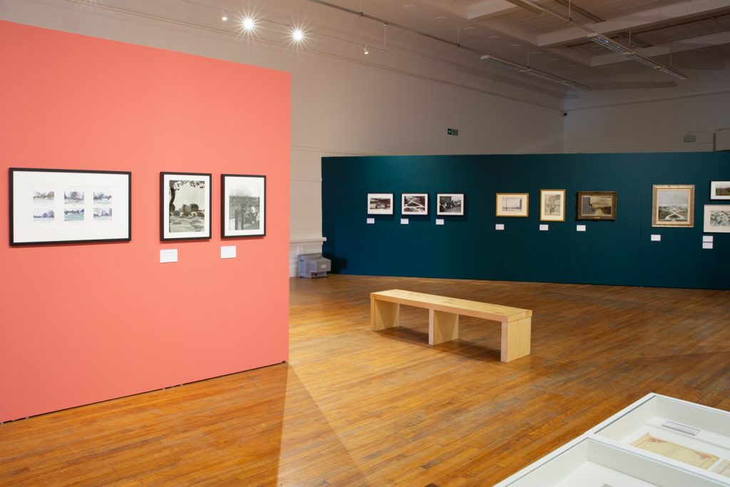 A landscape image showing the You Belong Here exhibition installed at Salford Museum and Art Gallery. On the left, a coral pink diving wall stands, with three artworks focusing on the green spaces of Salford hung. Behind and at an angle to the coral wall, stands a deep green wall with 9 more work’s focusing on Salford’s green spaces hung. Between them on the gold toned wood floor sits a gallery bench.
