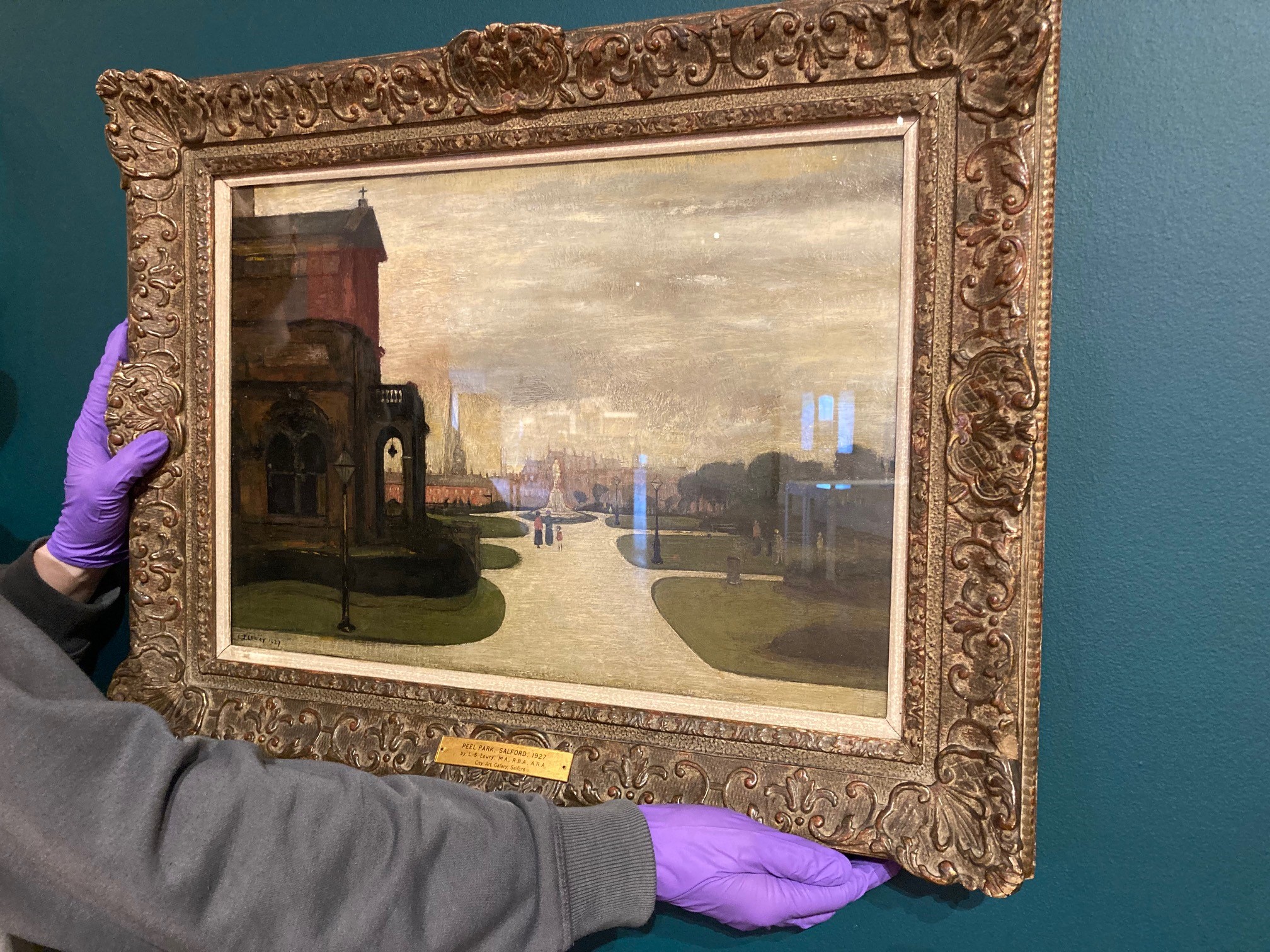 A landscape painting in an ornate gold frame, being held against a green wall. The technicians hands are visible, holding the work up using purple gloves. The image is a historic oil painting by LS Lowry, showing the early days of Peel  Park